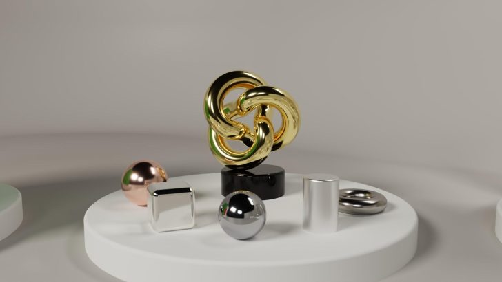 A 3D rendering features various reflective objects including a central interlocked gold structure, silver and rose gold spheres, a cuboid, and cylinders on round white platforms.